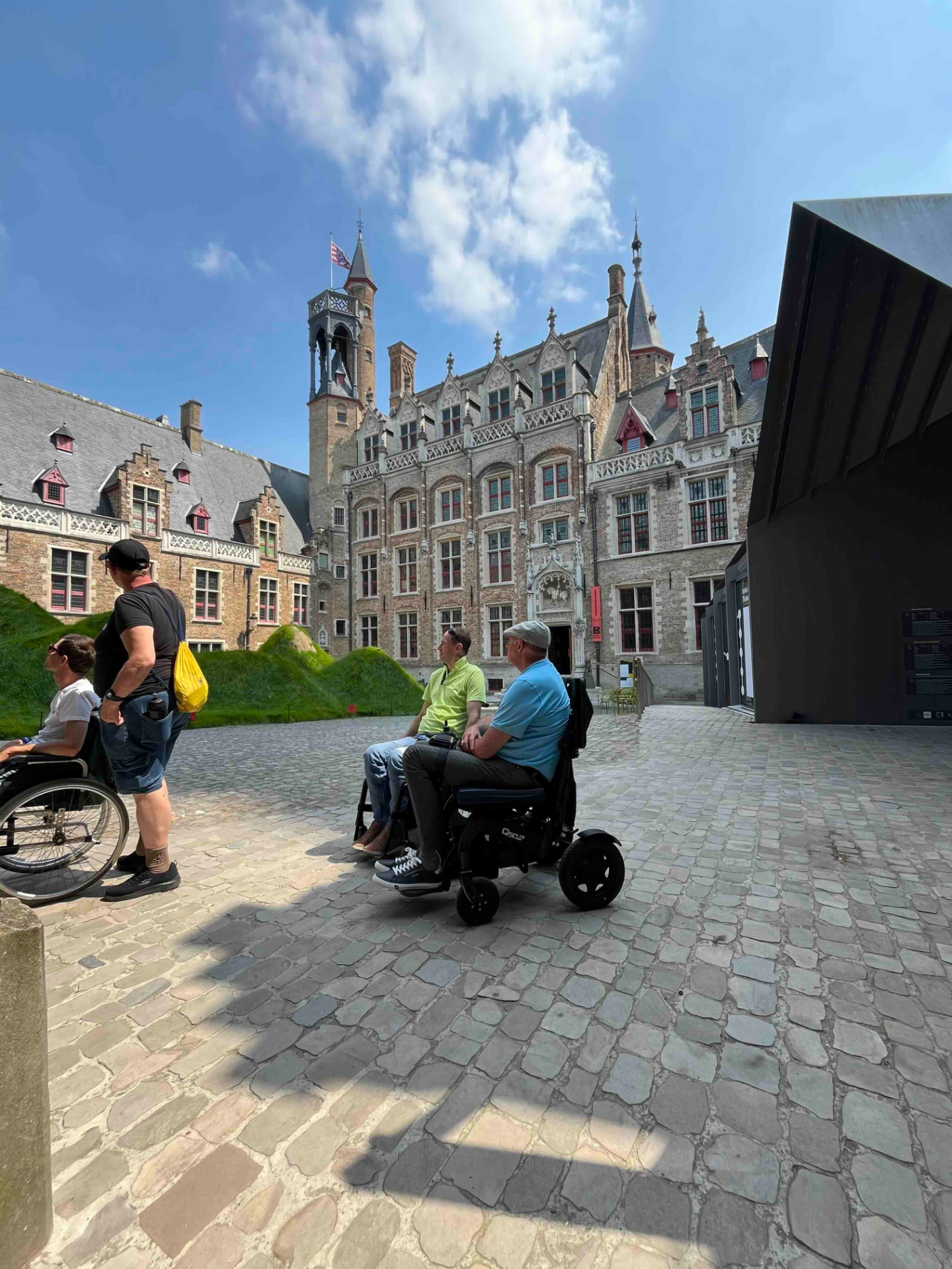 Des visiteurs en fauteuil roulant découvrent ensemble une place pavée historique ensoleillée.